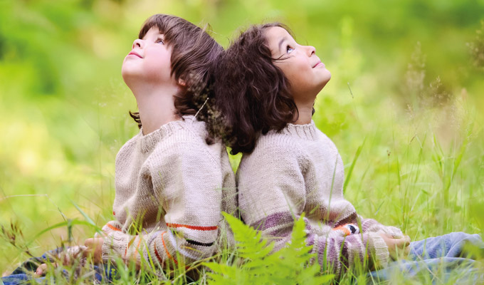 Bild zeigt zwei Kinder, die Rücken an Rücken im Gras sitzen und nach oben schauen